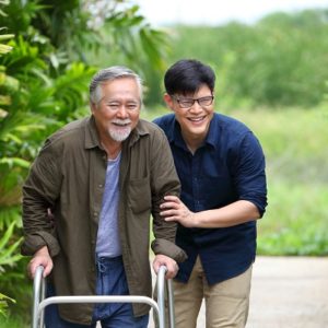 Happy elder Asian man using walker while walking for exercise around the garden with his son walks with him
