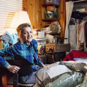Older adult woman sitting in a messy cluttered room bedroom looking at camera with serious expression