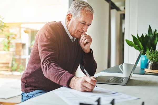 Older-adult man sitting at desk, pen in hand, at computer, contemplating decisions